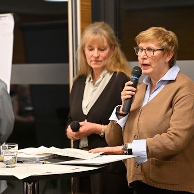 Drei Personen stehen an einem Stehtisch und leiten eine Versammlung.