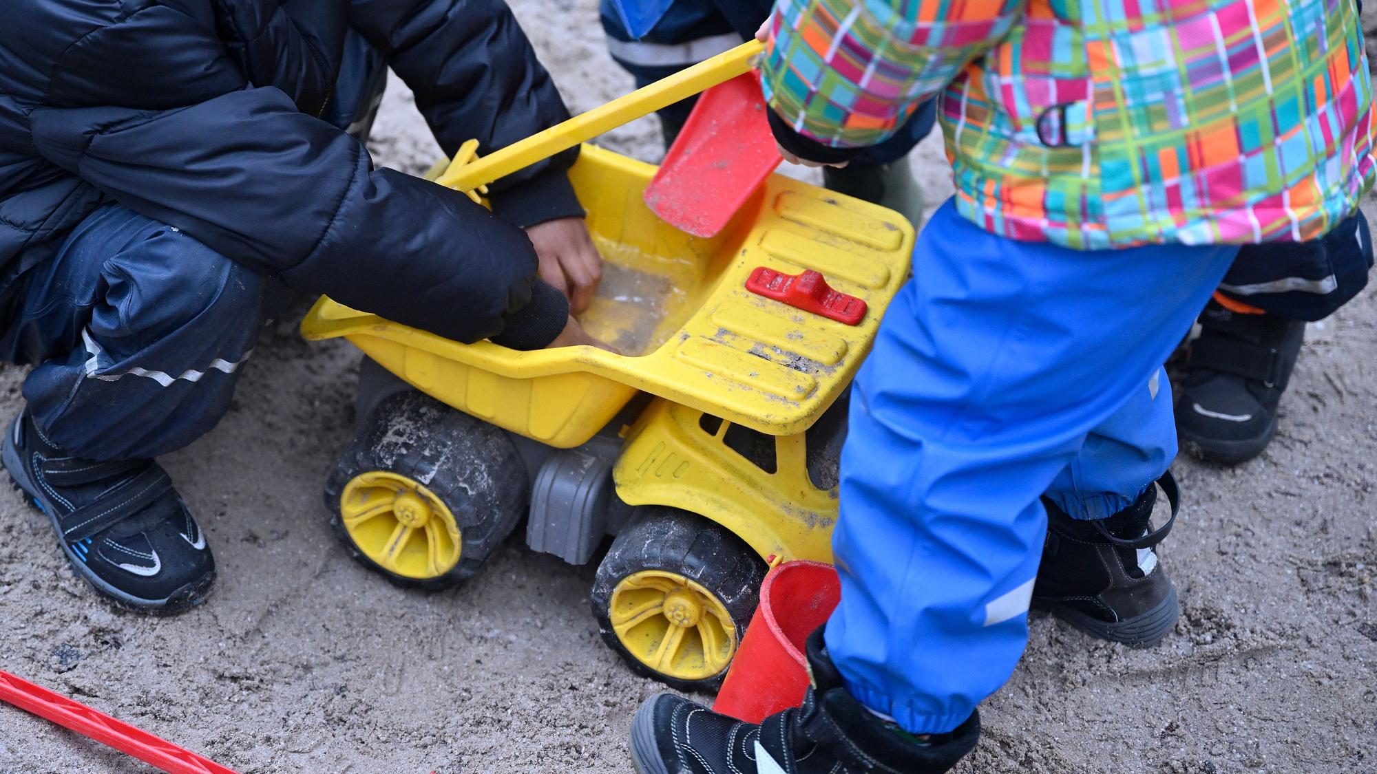 Kinder spielen in einem Sandkasten mit einem gelben Kipplader aus Plastik.