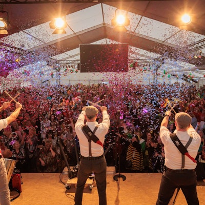 Auf einer Bühne stehen drei Musiker, die Drummer-Sticks in den Händen halten und diese vor den Gesichtern kreuzen. Vor der Bühne jubeln zahlreiche verkleidete Frauen. Von der Decke regnet Konfetti.