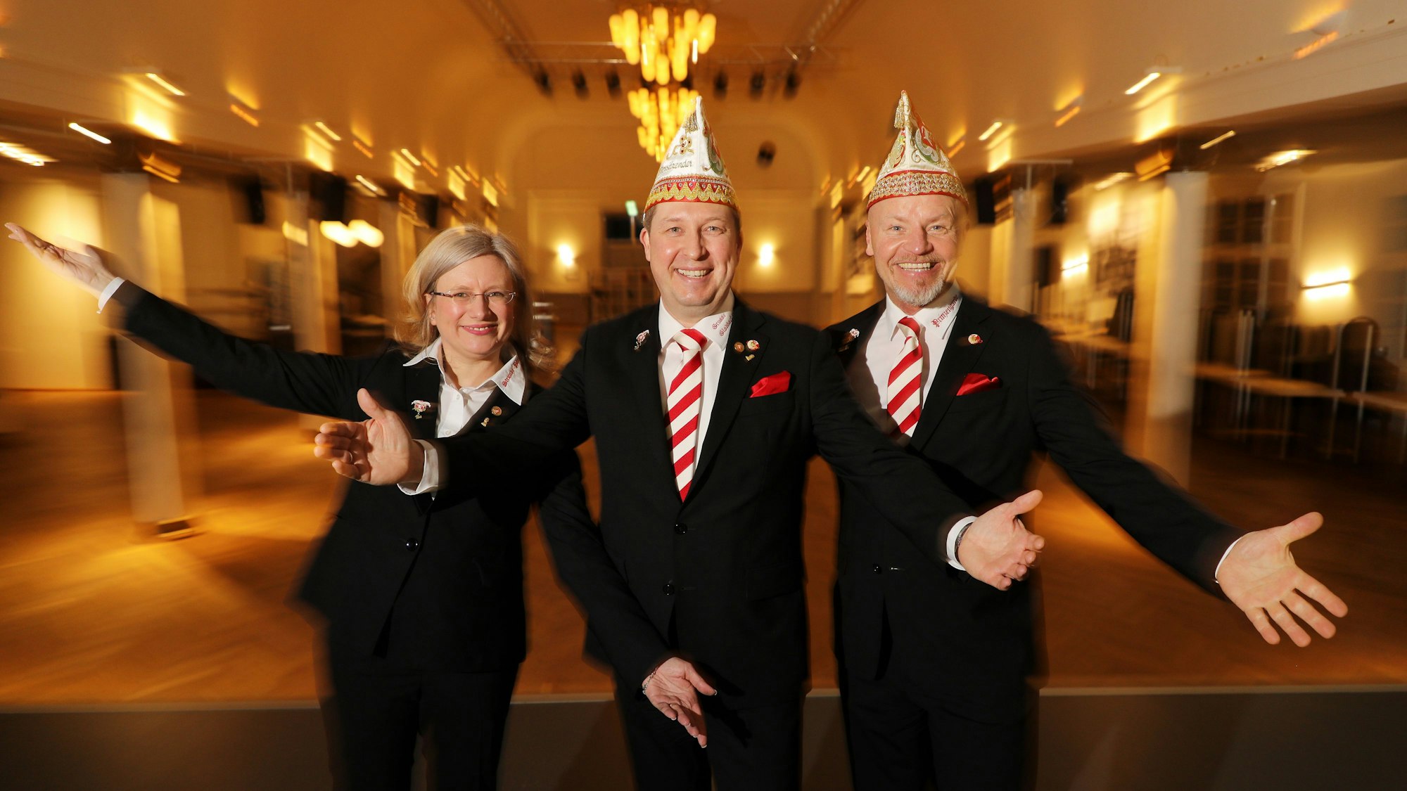 Das Dreigestirn steht in schwarzen Anzügen in einem Saal und hält die Hände nach vorne.