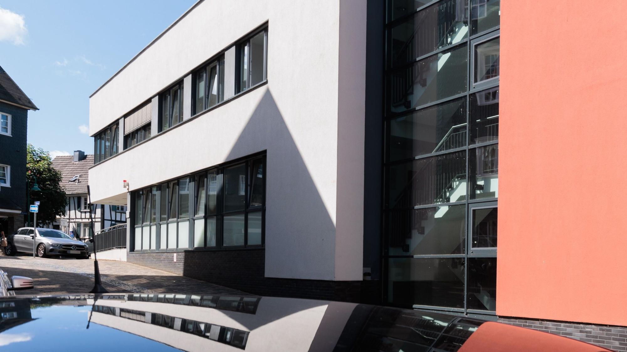 Ein modernes weißes Gebäude mit schwarzen Fensterrahmen; ein Anbau ist rot gestrichen.Das Haus spiegelt sich im Dach eines Autos.