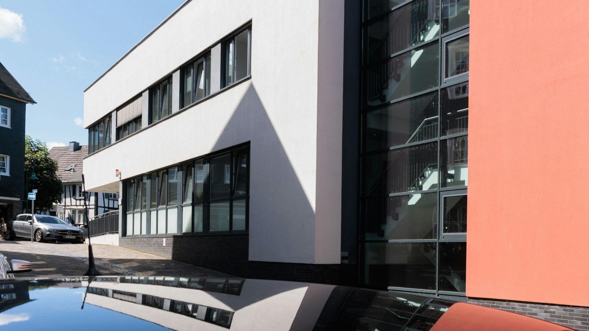 Ein modernes weißes Gebäude mit schwarzen Fensterrahmen; ein Anbau ist rot gestrichen.Das Haus spiegelt sich im Dach eines Autos.