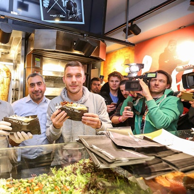 Lukas Podolski posiert mit Kollegen bei der Eröffnung eines Mangal-Döner-Ladens. Die Presse und Fans stehen im Hintergrund.