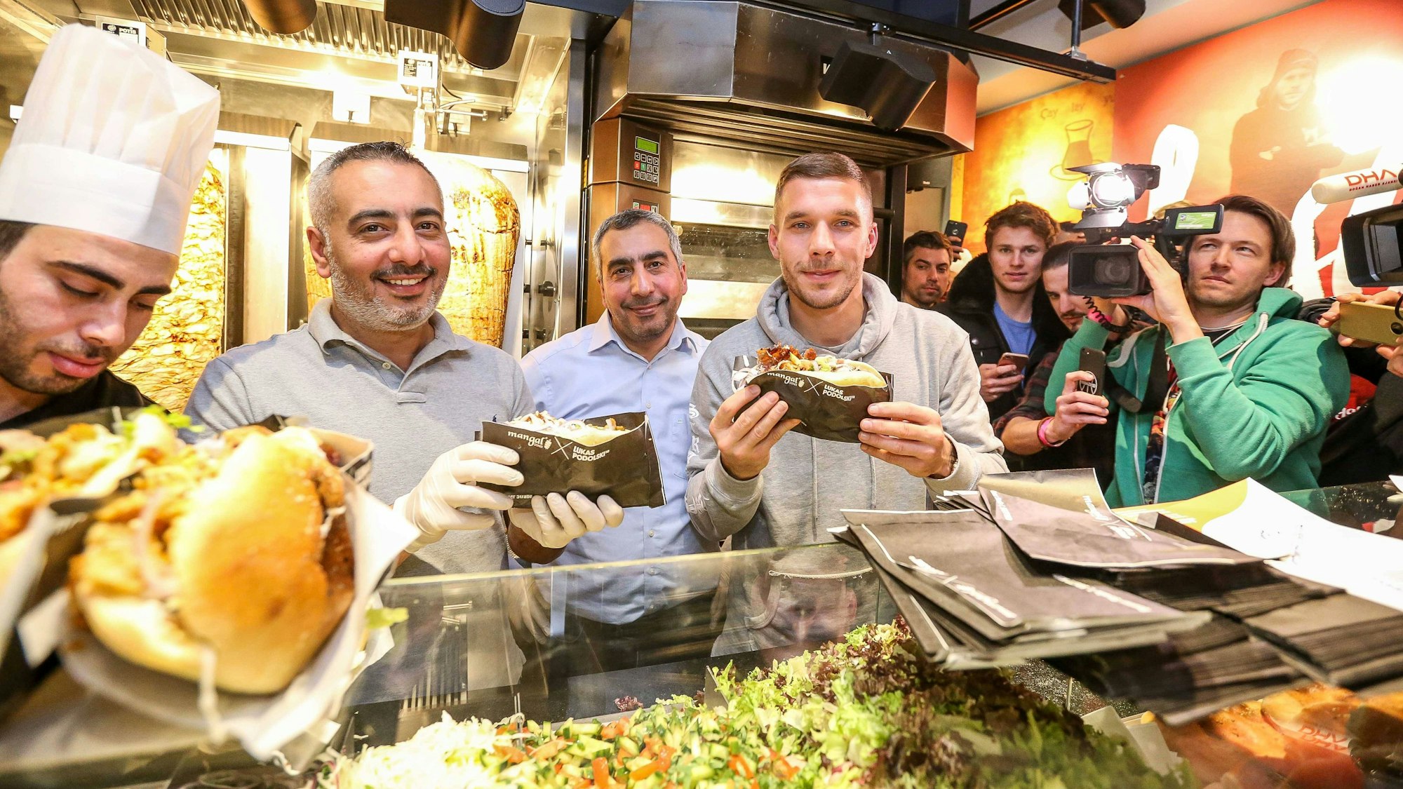 Lukas Podolski posiert mit Kollegen bei der Eröffnung eines Mangal-Döner-Ladens. Die Presse und Fans stehen im Hintergrund.