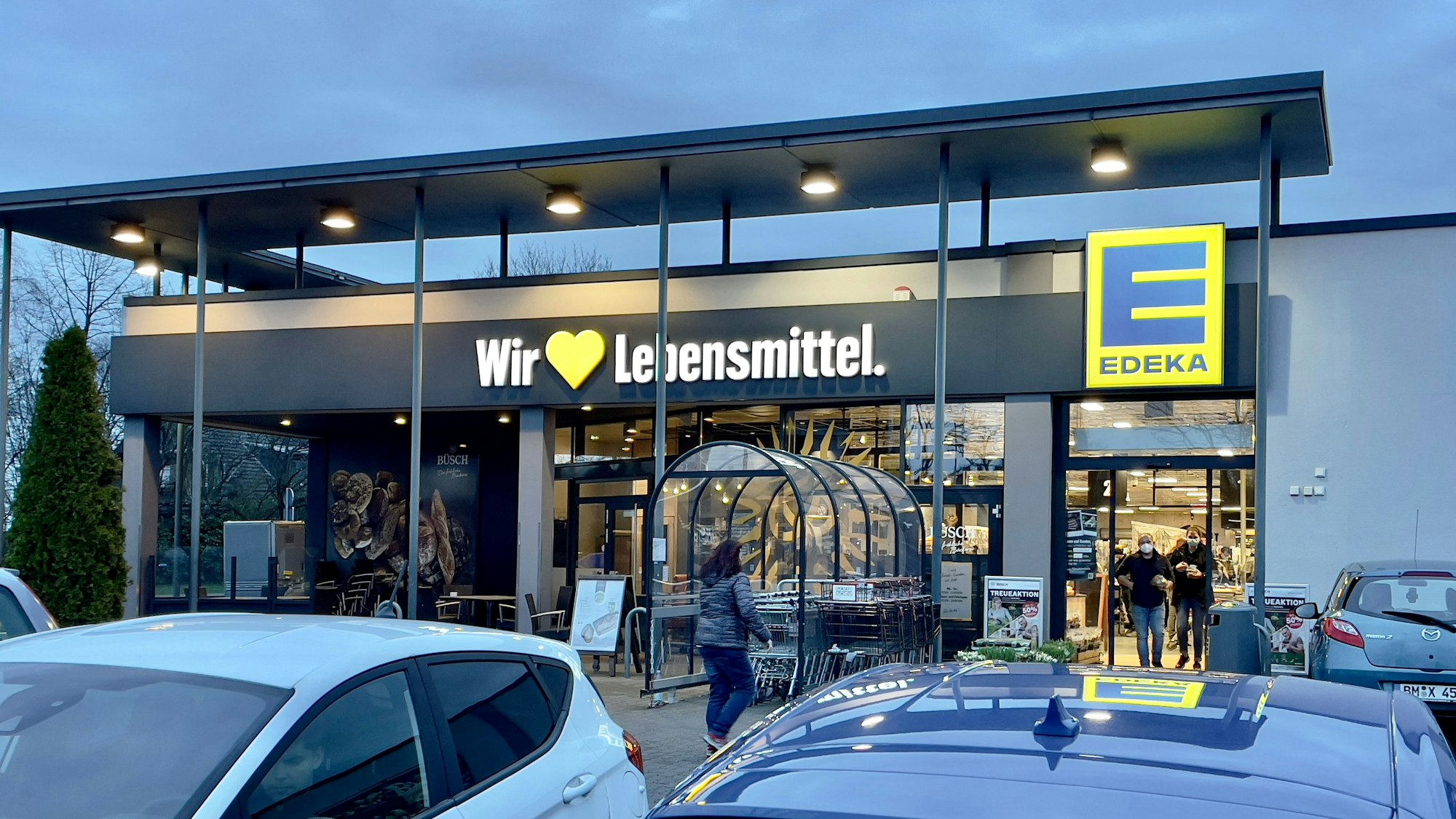 Der Eingang einer Edeka-Filiale mit parkenden Autos im Vordergrund.