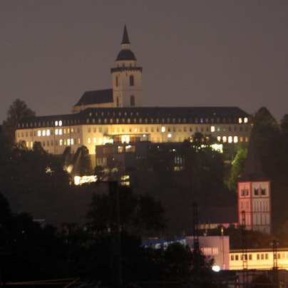 Vor dem nächtlichen Himmel hebt sich die erleuchtete Abtei auf dem Michaelsberg ab, der Turm der Kirche ragt in die Höhe. Weiter unten steht die rot-weiße Servatiuskirche.