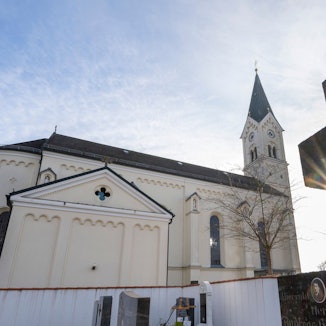 Vor dem weißen Kirchengebäude St. Nikolaus in Garching an der Alz wird ein Kreuz von der Sonne angestrahlt.