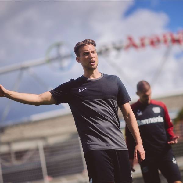 Dennis Eck (vorne) steht vor der BayArena und gibt mit erhobenem Arm Anweisungen, Fußball-Profi Robert Andrich von Bayer 04 Leverkusen schaut auf seine linke Hand.