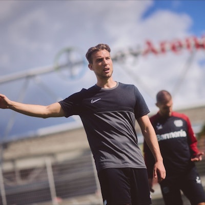 Dennis Eck (vorne) steht vor der BayArena und gibt mit erhobenem Arm Anweisungen, Fußball-Profi Robert Andrich von Bayer 04 Leverkusen schaut auf seine linke Hand.