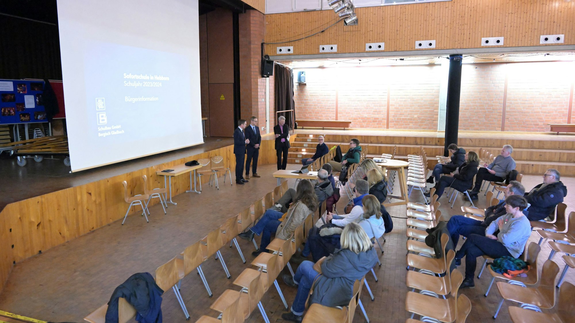 Ein gutes Dutzend Menschen sitzen in einer Schulaula und hören der Präsentation dreier Männer vorne zu.