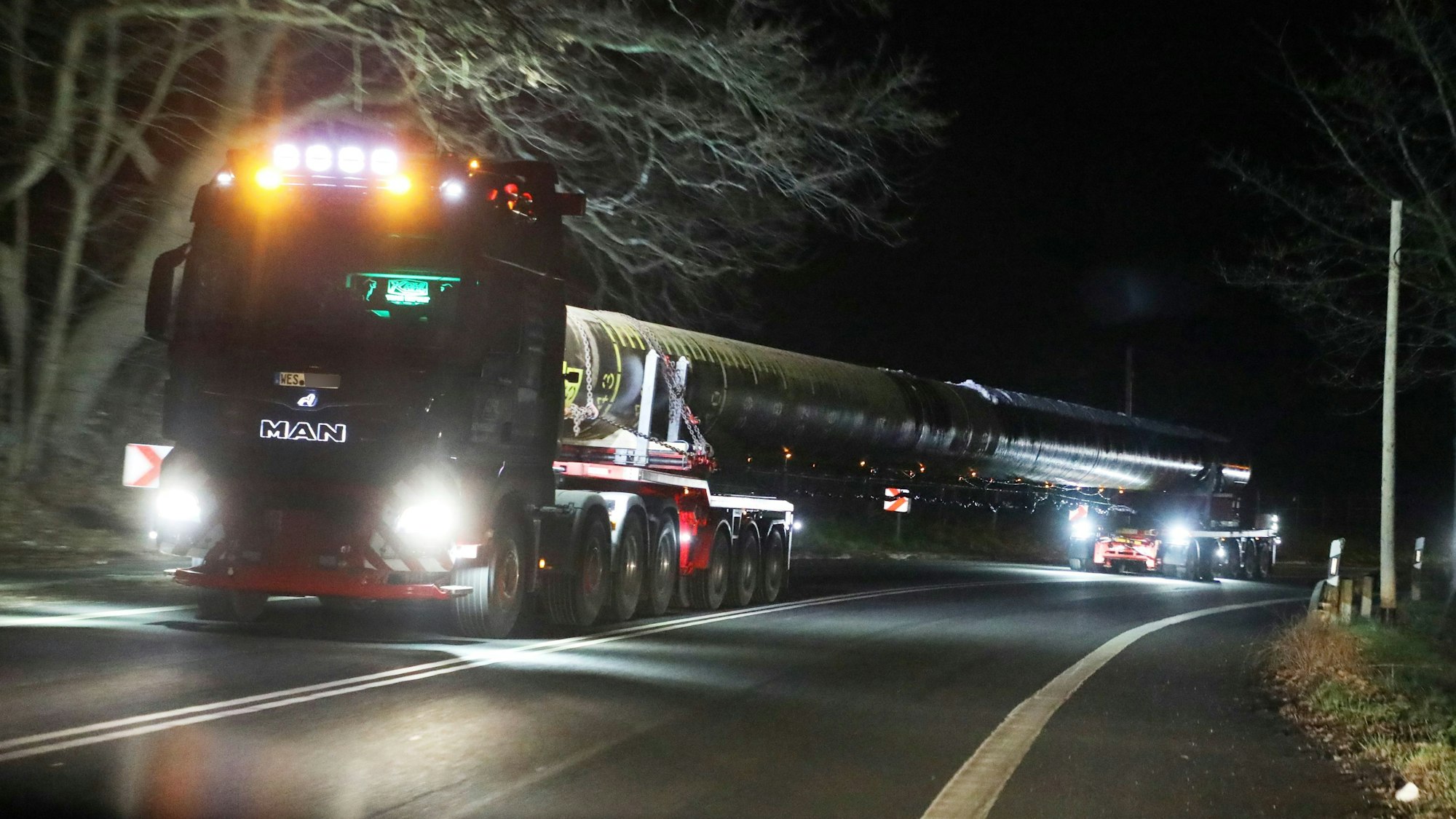 Ein Schwertransport mit gelbem Warnlicht fährt auf einer Landstraße. Er hat ein langes Rohr geladen.