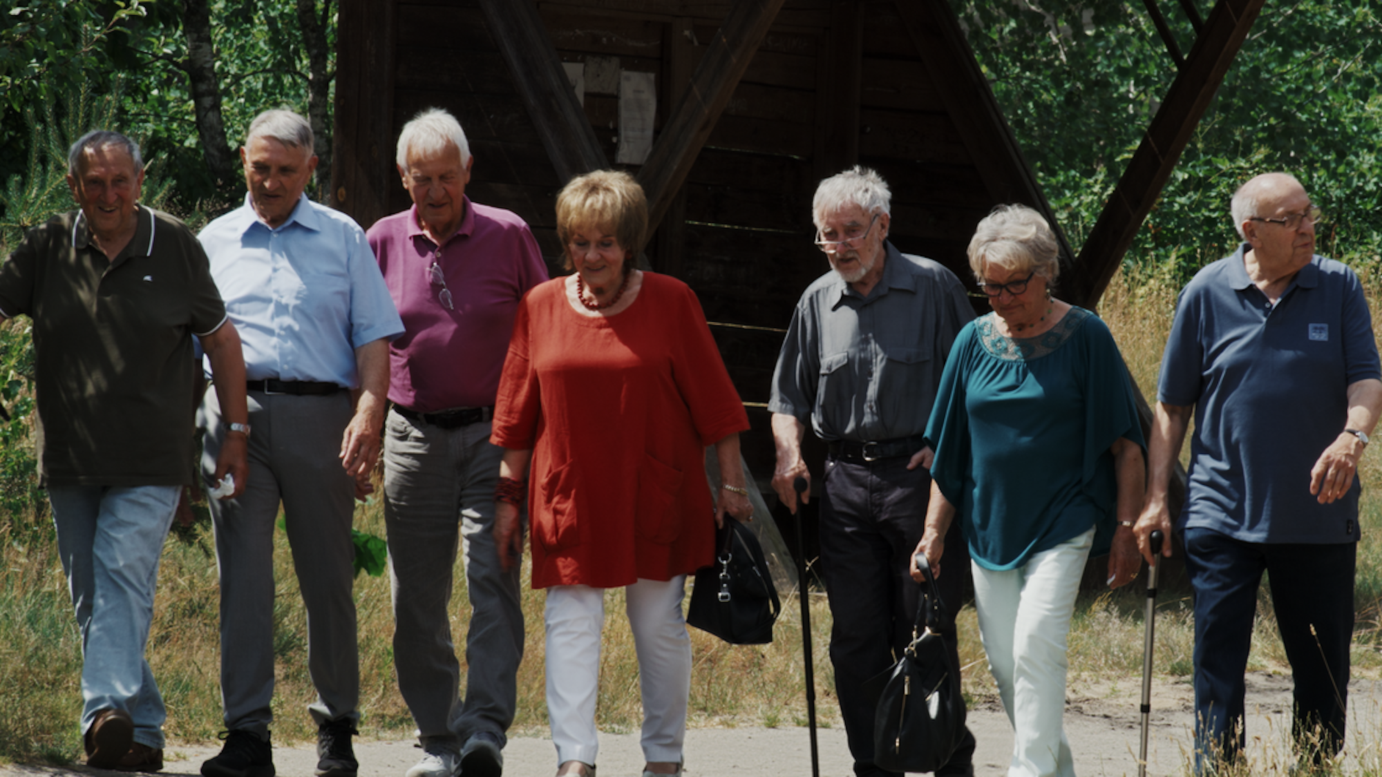 Sechs ältere Männer und zwei Frauen gehen nebeneinander teils mit Hilfe eines Stocks auf einem Waldweg.