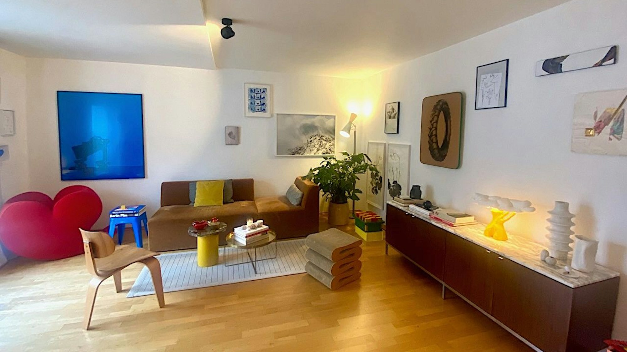 Blick in ein Wohnzimmer, in dem ein blaues Bild, ein Herz-Stuhl und eine gelbe Lampe und Kissen zu sehen sind.