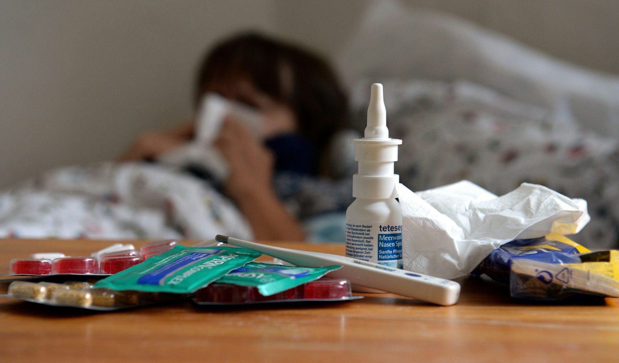Verschiedene Erkältungsmittel liegen auf einem Tisch. Im Hintergrund liegt jemand im Bett.