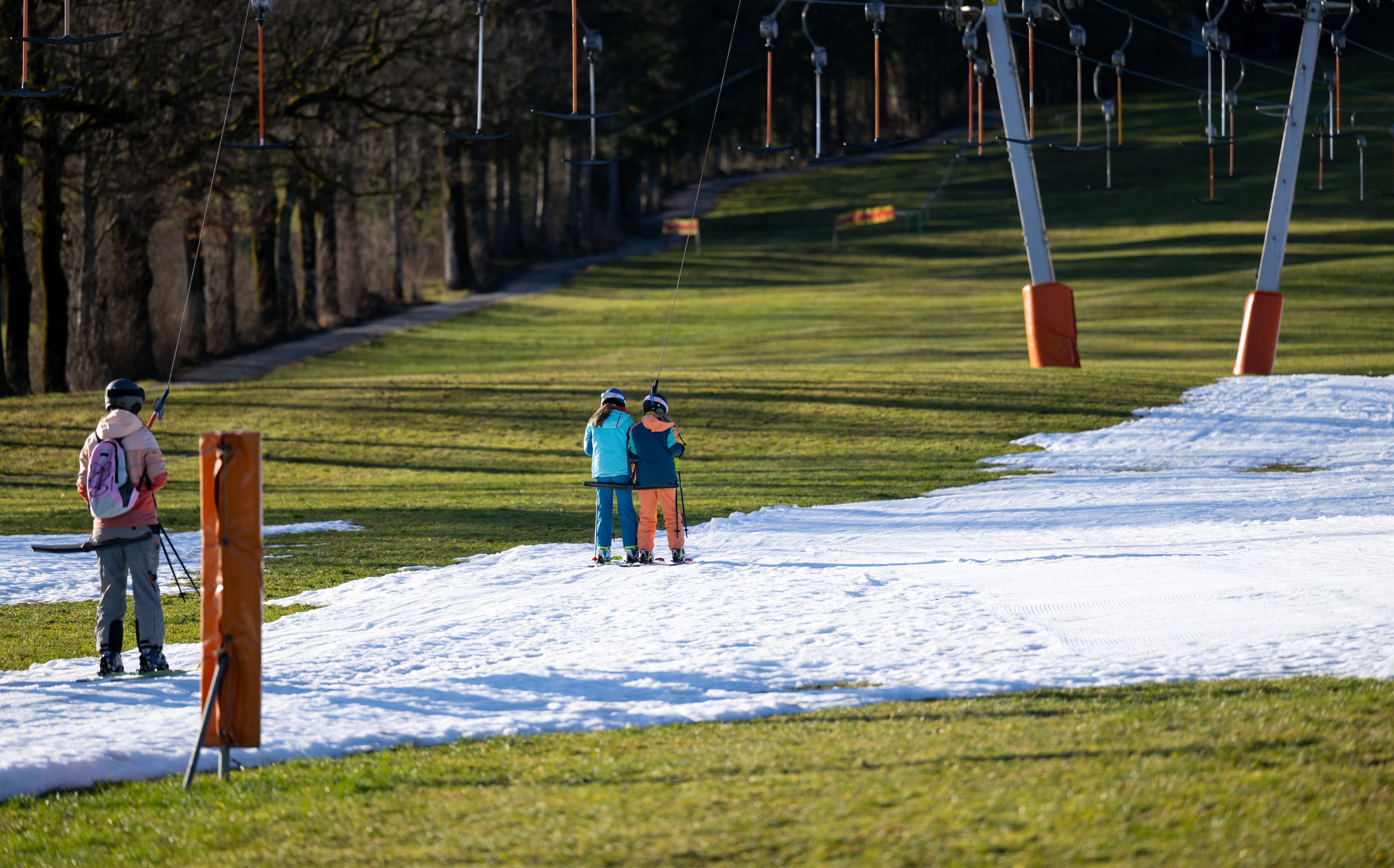 Wintersportler fahren in einem bayrischen Skigebiet zwischen grünen Wiesen mit einem Schlepplift die beschneite Piste hinauf. Auch die Natur leidet unter der ungewöhnlichen Wärme.