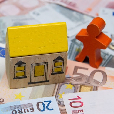 Man sieht ein Bauklotz-Haus, das auf Geldscheinen steht. Rechts neben dem Haus steht eine Holzfigur, links ein Holzbaum.