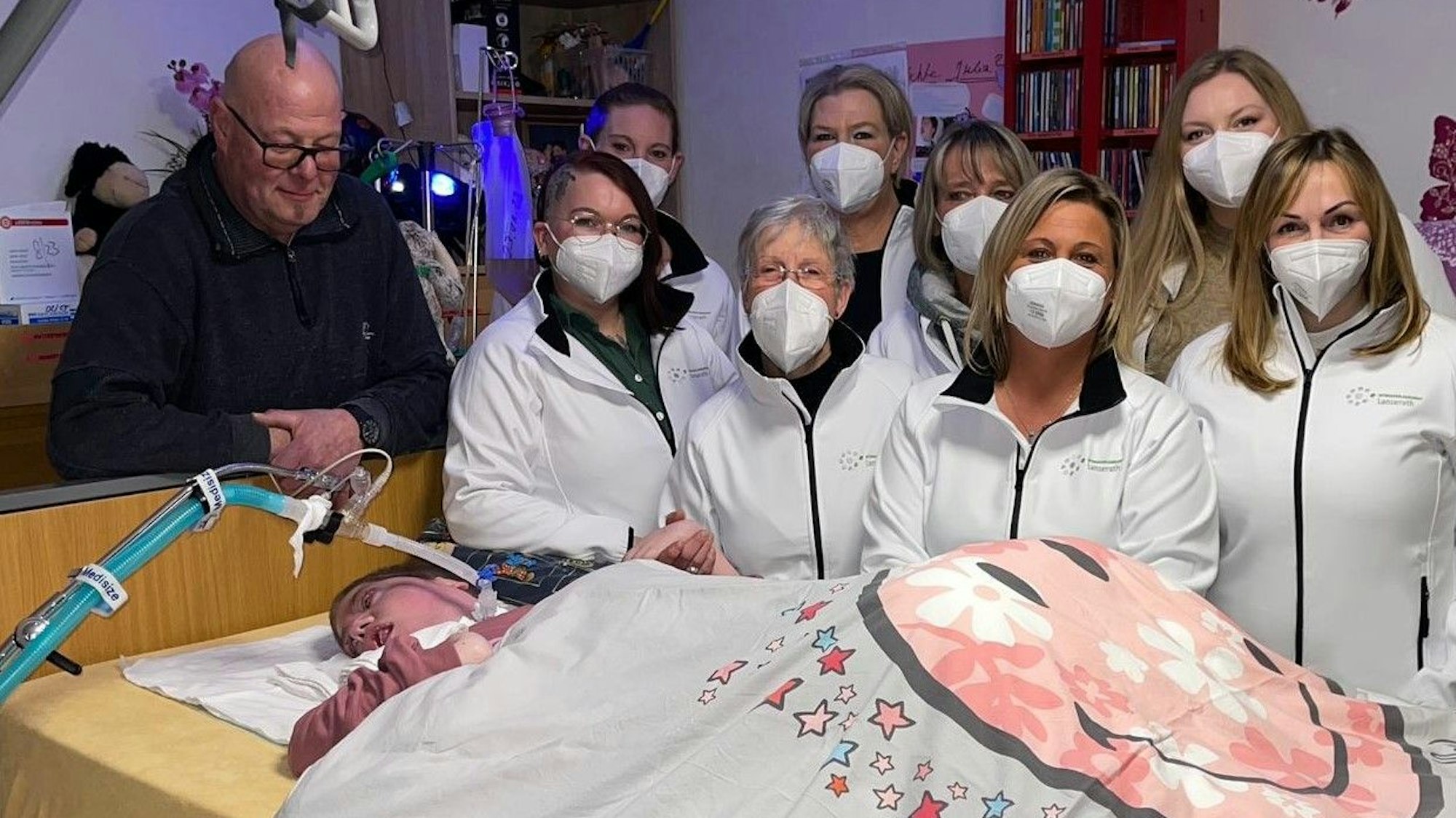 Man sieht Julia in einem Pflegebett liegen, am Kopfende ihr Vater, daneben acht Personen des Pflegeteams.