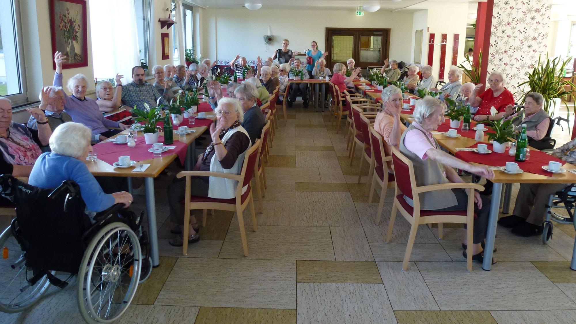 Man sieht einen Raum, dem Senioren an einem großen, u-förmigen Tisch sitzen, einige winken in die Kamera. Am Kopfende stehen zwei Personen.