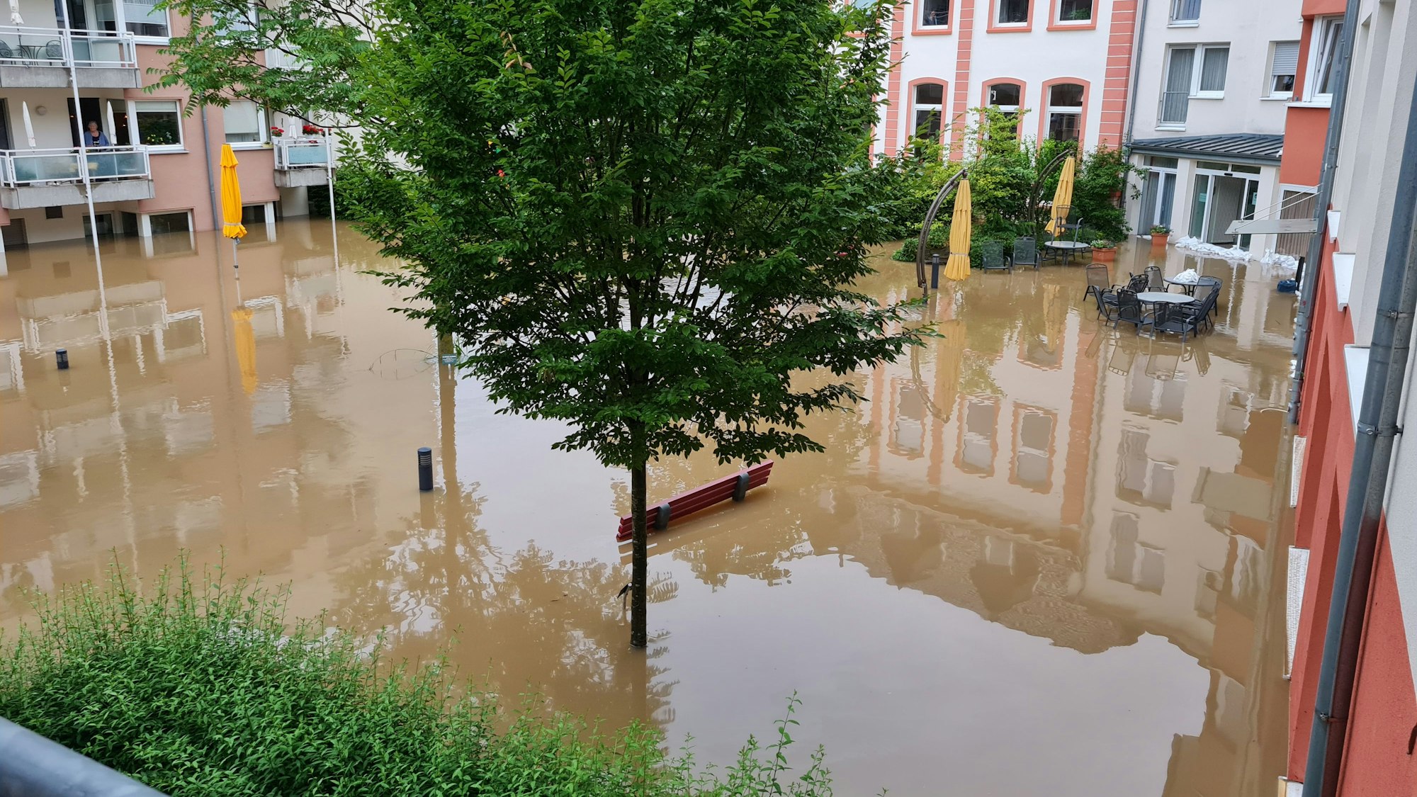 Man sieht einen Innenhof, der überflutet wurde. Es stehen Bäume im Wasser, Bänke und Tische schauen aus dem Wasser heraus.