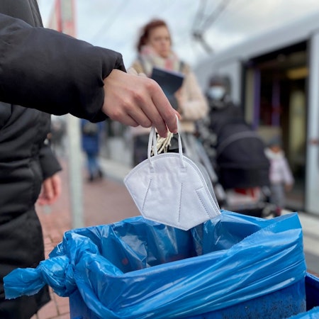 Auf dem Foto sind eine Hand und eine Maske zu sehen. Die Maske wird in den Mülleimer befördert.