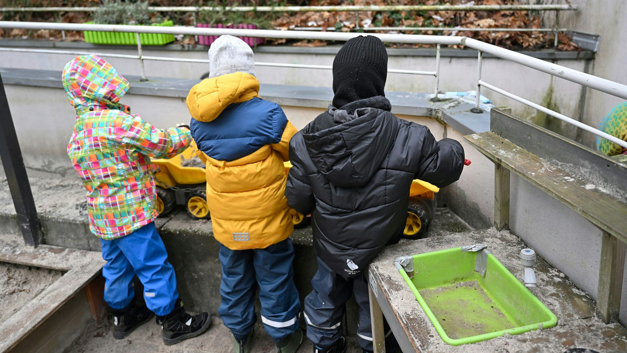 Man sieht drei Kinder, die auf einem Balkon spielen. Sie tragen Wintersachen und stehen mit dem Rücken zur Kamera.