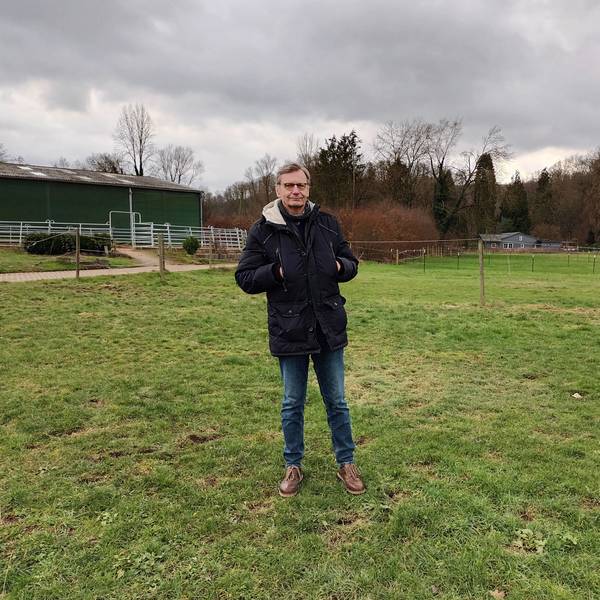 Ein Mann im Anorak steht auf einer grünen Wiese. Im Hintergrund steht ein Pferdestall.