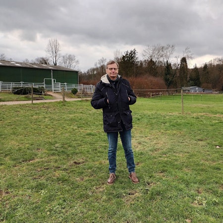 Ein Mann im Anorak steht auf einer grünen Wiese. Im Hintergrund steht ein Pferdestall.