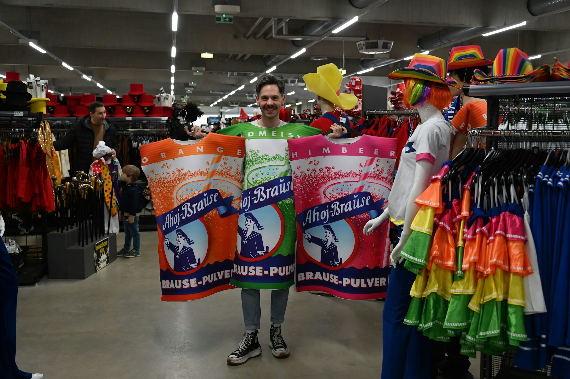 Ein Mann ist als grüne Ahoj-Brause verkleidet und hält zwei weitere Ahoj-Brause-Kostüme in einem Karnevalsladen hoch.