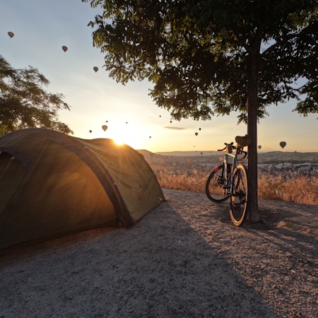 Ein Rad lehnt an einen Baum neben einem Zelt im Hintergrund geht die Sonne unter und bescheint Heißluftbaloons am Himmel
