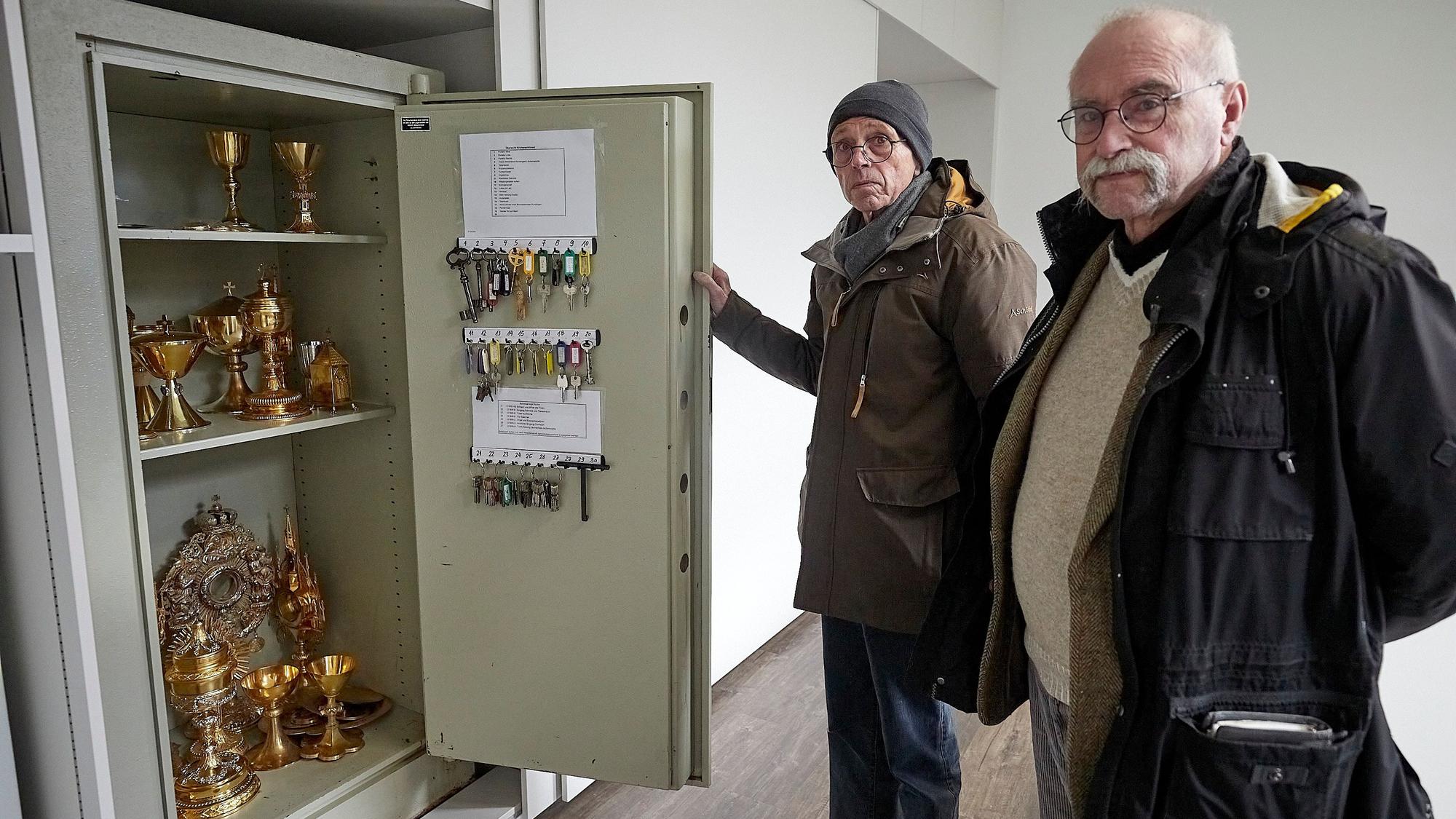 Zwei Männer stehen an einem Tresor, in dem sich goldene Sakralgefäße befinden.