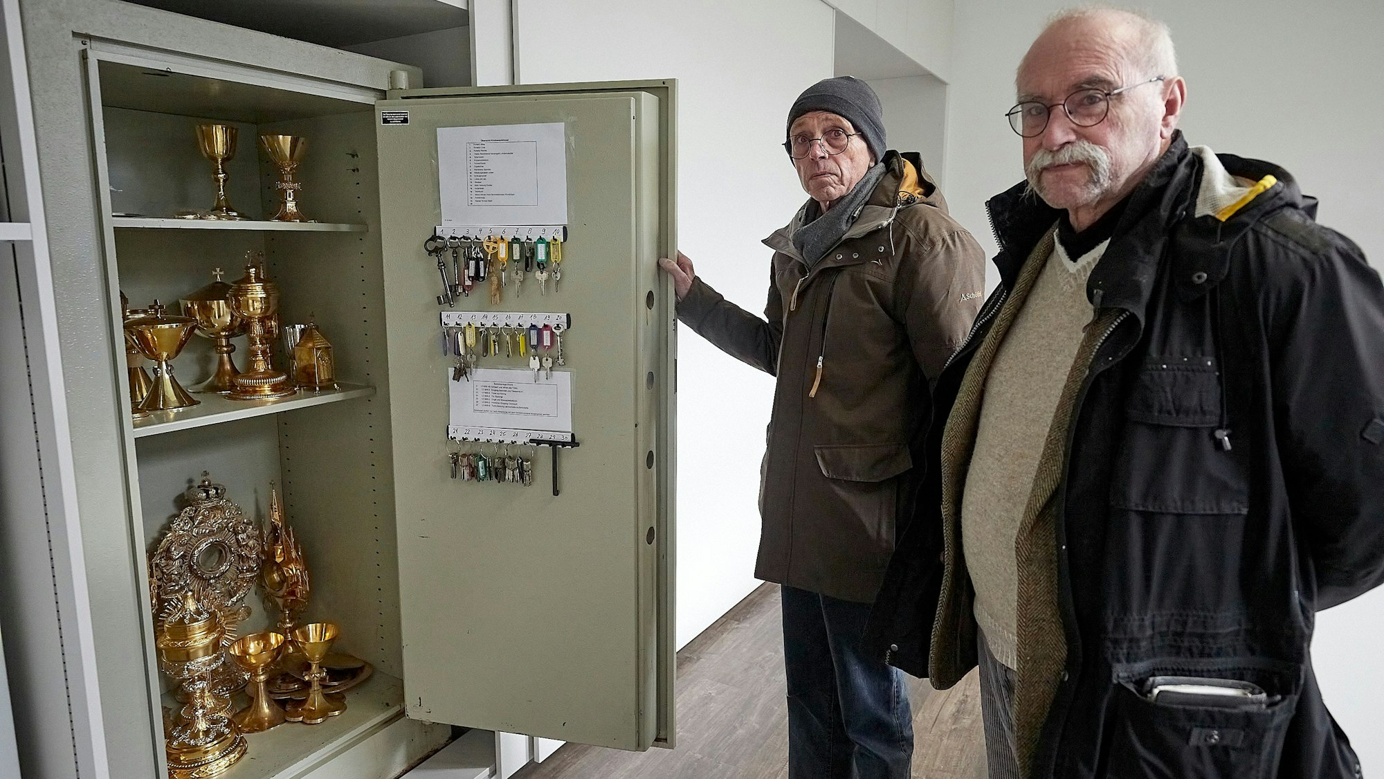Zwei Männer stehen an einem Tresor, in dem sich goldene Sakralgefäße befinden.