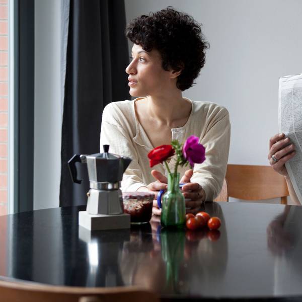 Eine Frau und ein Mann sitzen an einem Tisch. Die Frau schaut aus dem Fenster, der Mann in eine Zeitung.