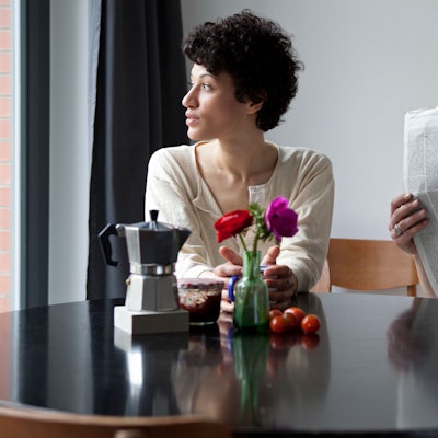 Eine Frau und ein Mann sitzen an einem Tisch. Die Frau schaut aus dem Fenster, der Mann in eine Zeitung.