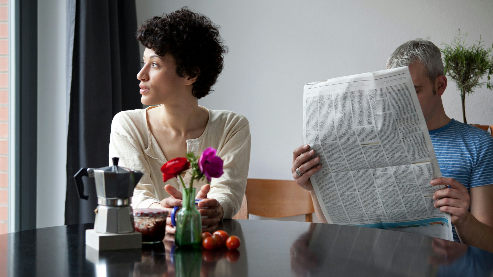 Eine Frau und ein Mann sitzen an einem Tisch. Die Frau schaut aus dem Fenster, der Mann in eine Zeitung.