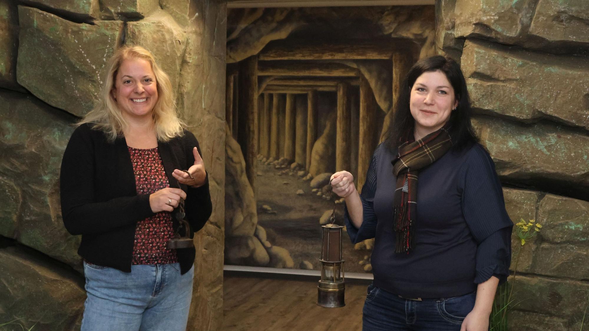 Katja Wonneberger-Kühr und Sarah-Sophie Riedel stehen vor einer künstlichen Steinwand, in der ein Stolleneingang aufgemalt ist.