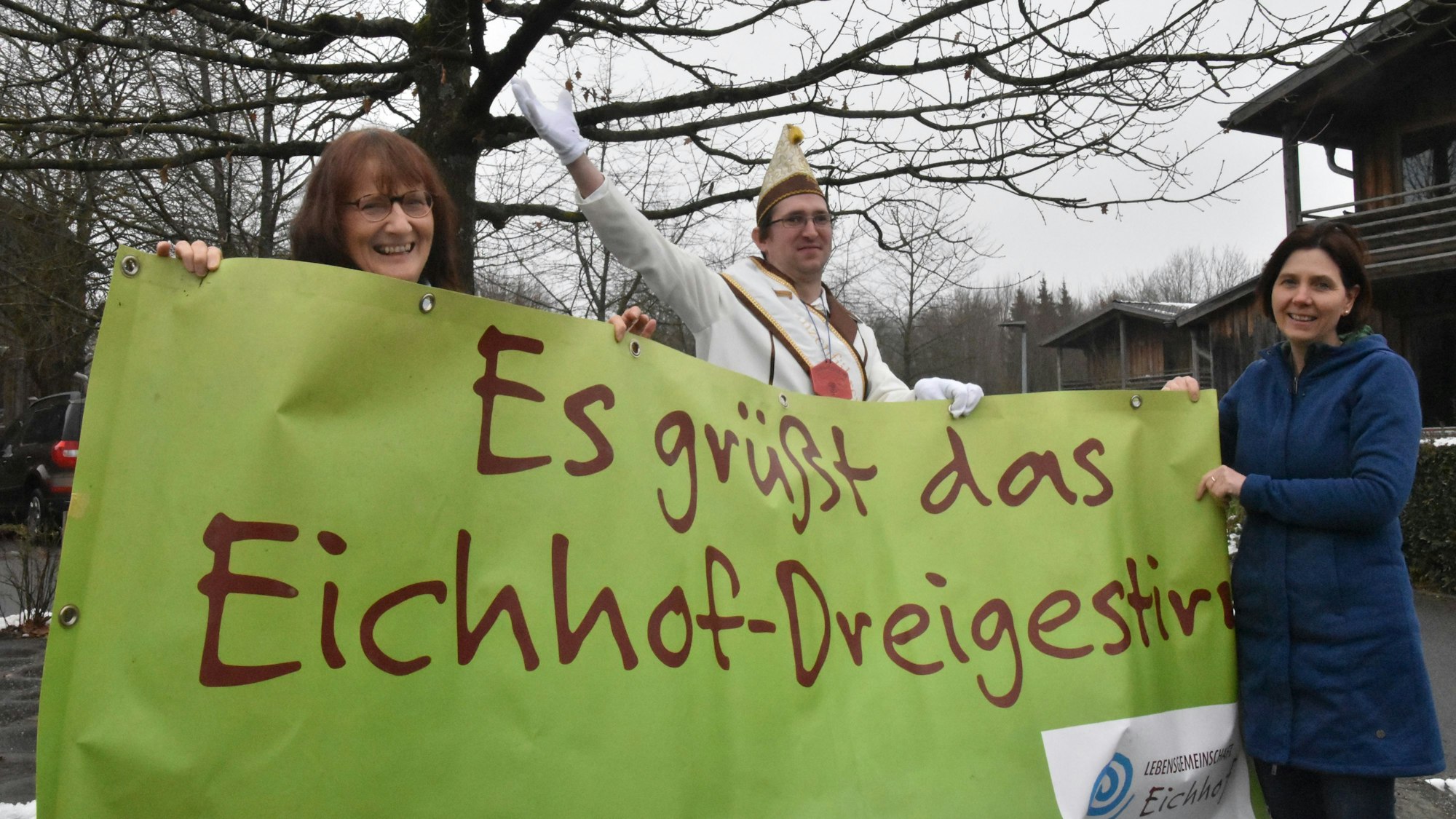 Drei Menschen haben ein grünes Banner in der Hand auf dem steht: „Es grüßt das Eichhof-Dreigestirn“.