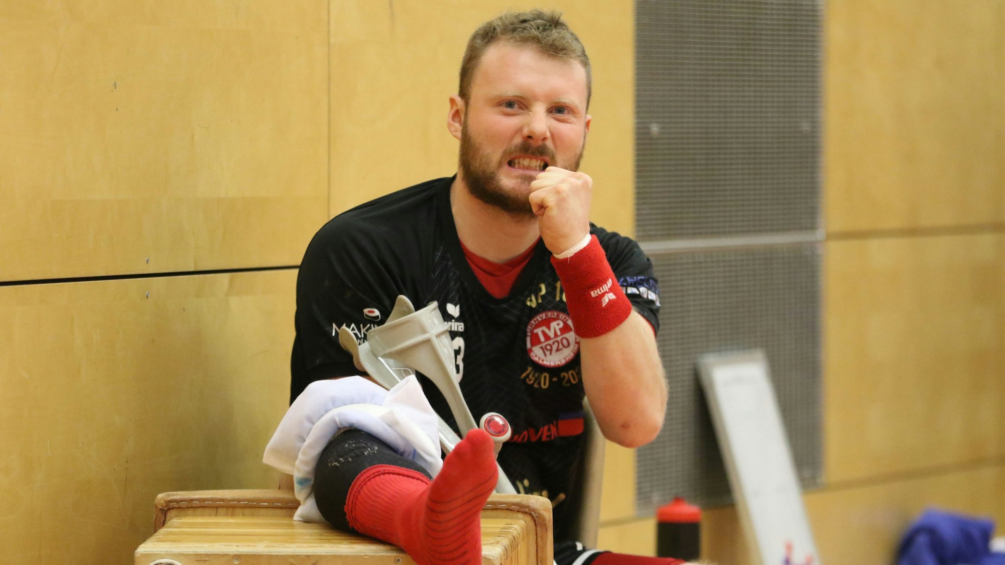 Der TVP-Spieler sitzt verletzt am Spielfeldrand, hat den Fuß hochgelegt und jubelt seiner Mannschaft zu.