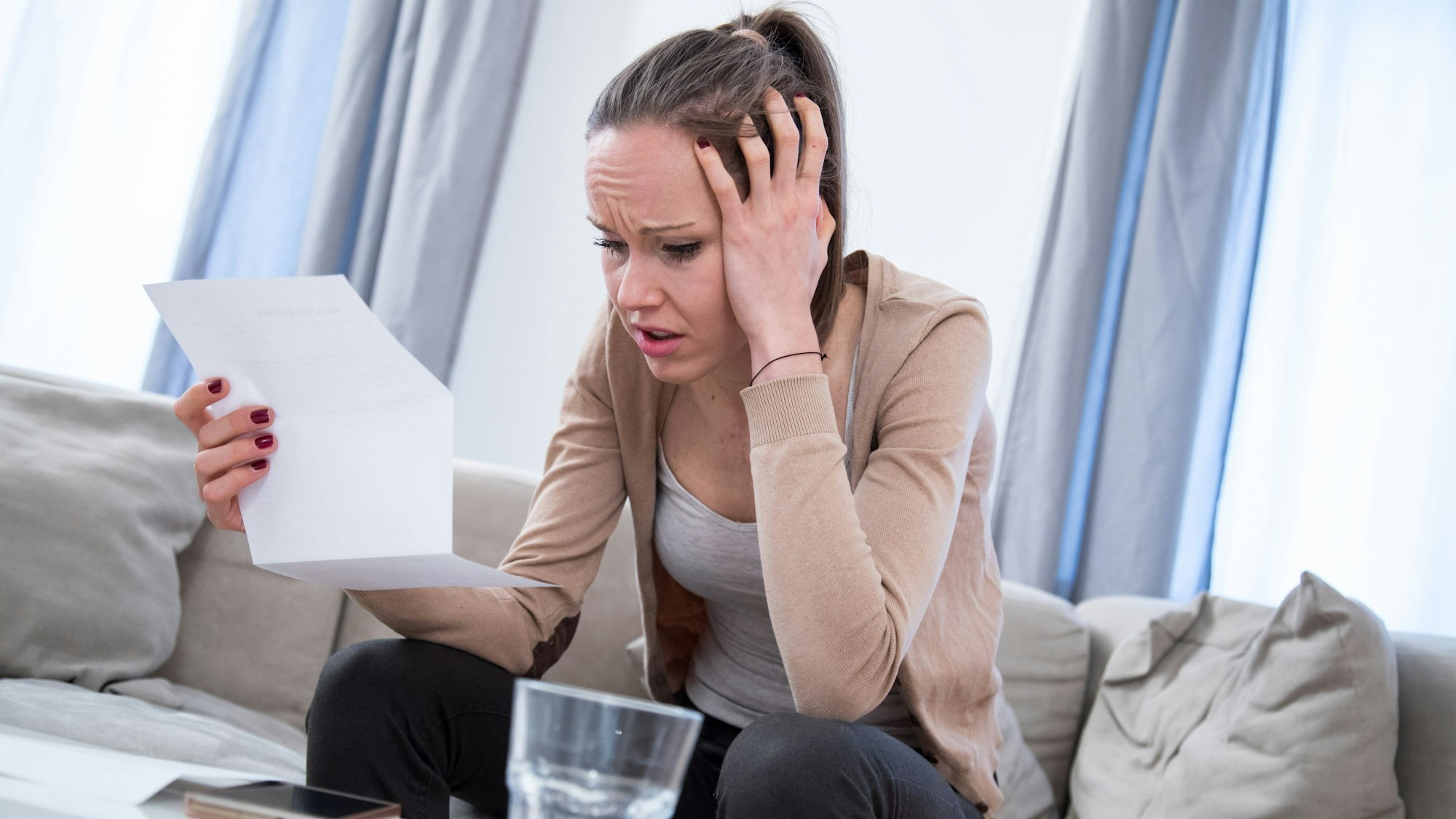 Eine Frau sitzt auf der Couch und schaut verzweifelt auf einen Brief, den sie in der Hand hält. (Symbolbild)