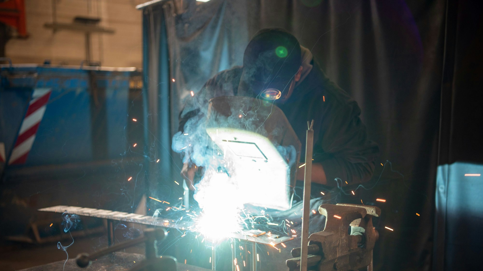 Hady Bah steht an einer Werkbank und schaut durch einen Sichtschutz, vor ihm fliegen helle Funken.