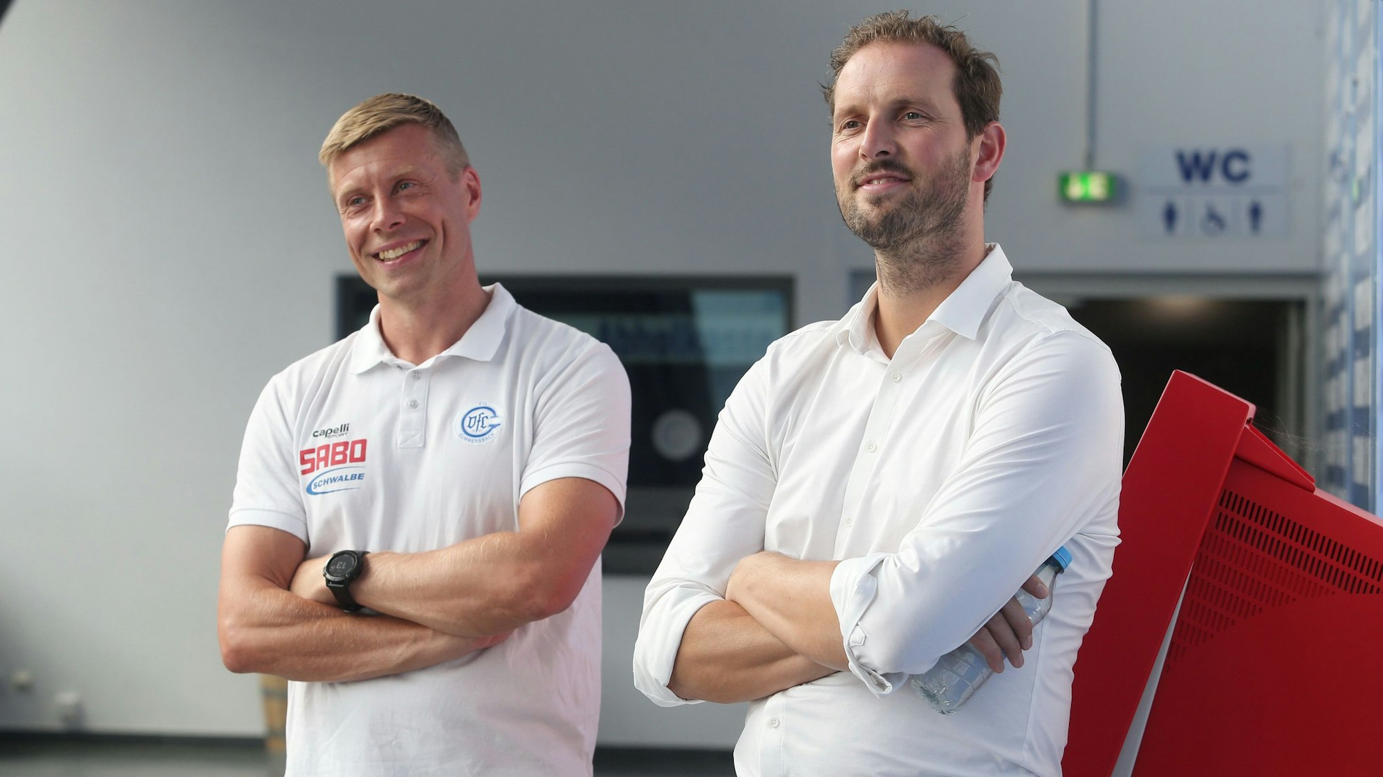 Zwei Männer in weißem Outfit stehen mit verschränkten Armen nebeneinander.