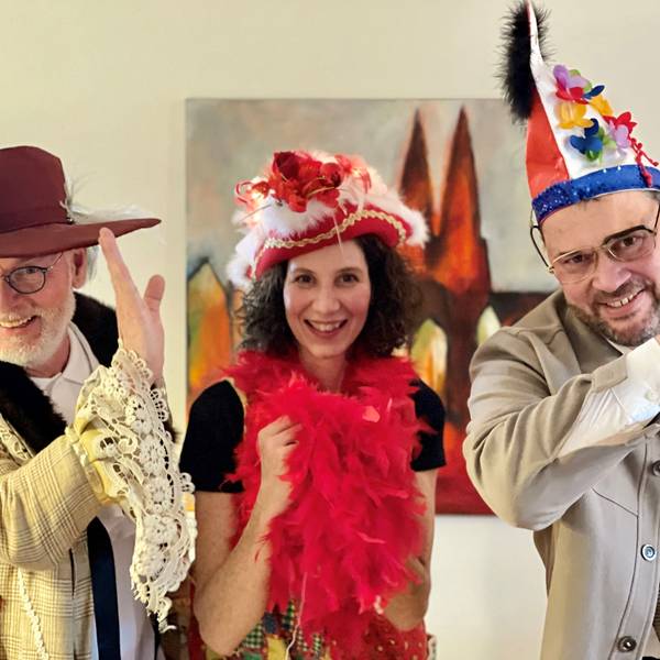 Ein Mann mit weißem Bart und Brille trägt Schlapphut und ein helle, karierte Jacke mit Rüschenärmel, eine dunkelhaarige Frau eine rote Federboa und einen roten Dreispitz, ein weiterer Mann eine blau-rote Narrenkappe. Sie heben die Hand an die Stirn zum jecken Gruß,