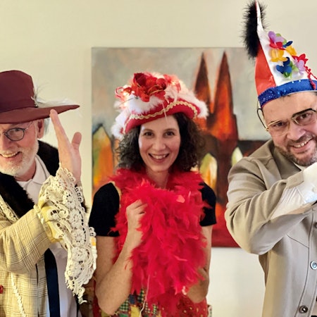 Ein Mann mit weißem Bart und Brille trägt Schlapphut und ein helle, karierte Jacke mit Rüschenärmel, eine dunkelhaarige Frau eine rote Federboa und einen roten Dreispitz, ein weiterer Mann eine blau-rote Narrenkappe. Sie heben die Hand an die Stirn zum jecken Gruß,