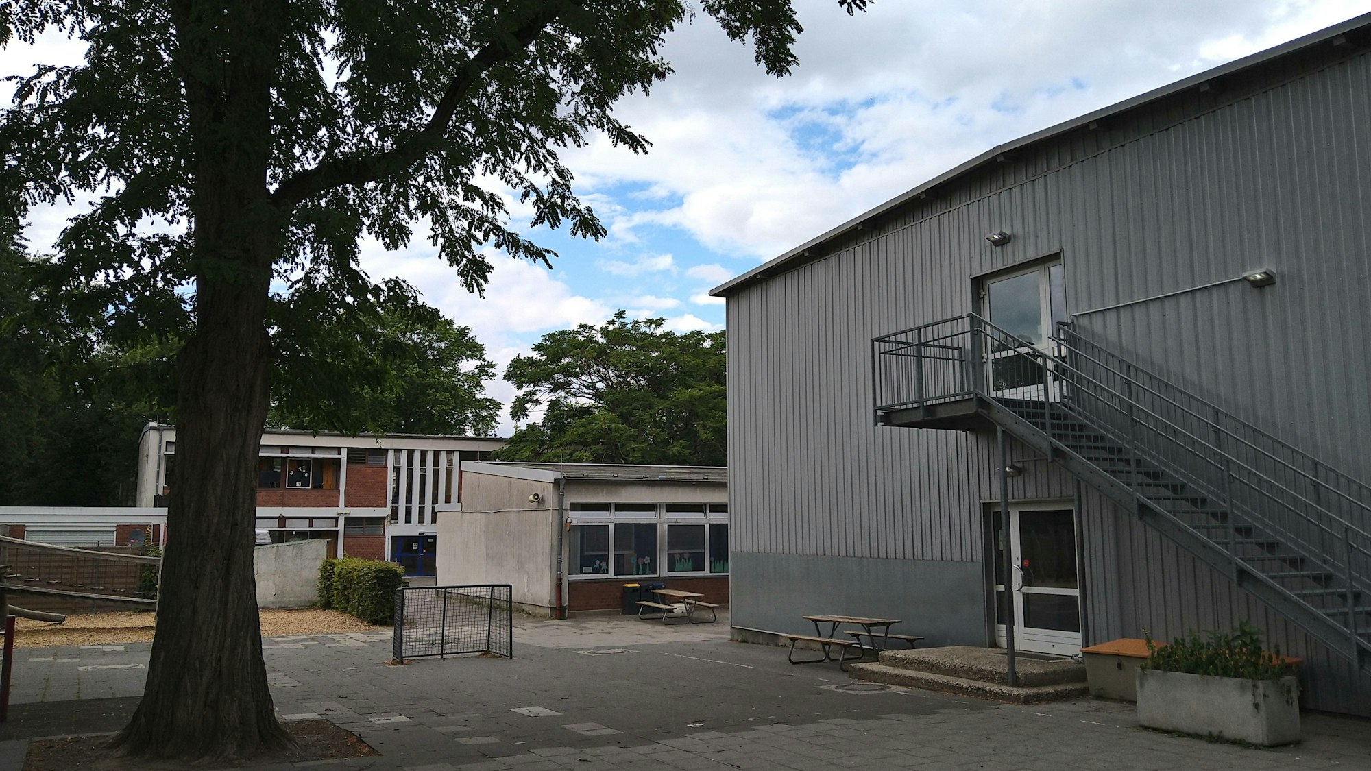 Auf einem Schulhof sind ein großer Baum und ein Container-Gebäude zu sehen, im Hintergrund zwei Flachbauten älteren Datums.