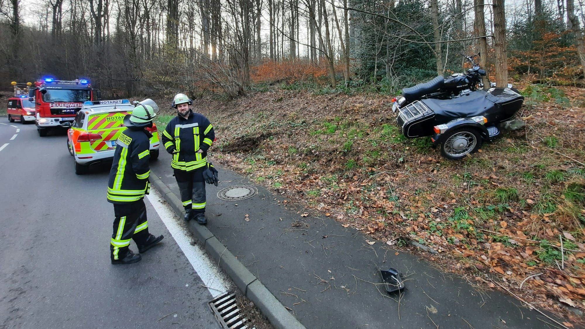 Rechts im Bild das Motorrad an einem Baum, links auf der Straße Feuerwehrleute, Notarztwagen und Feuerwehrautos.