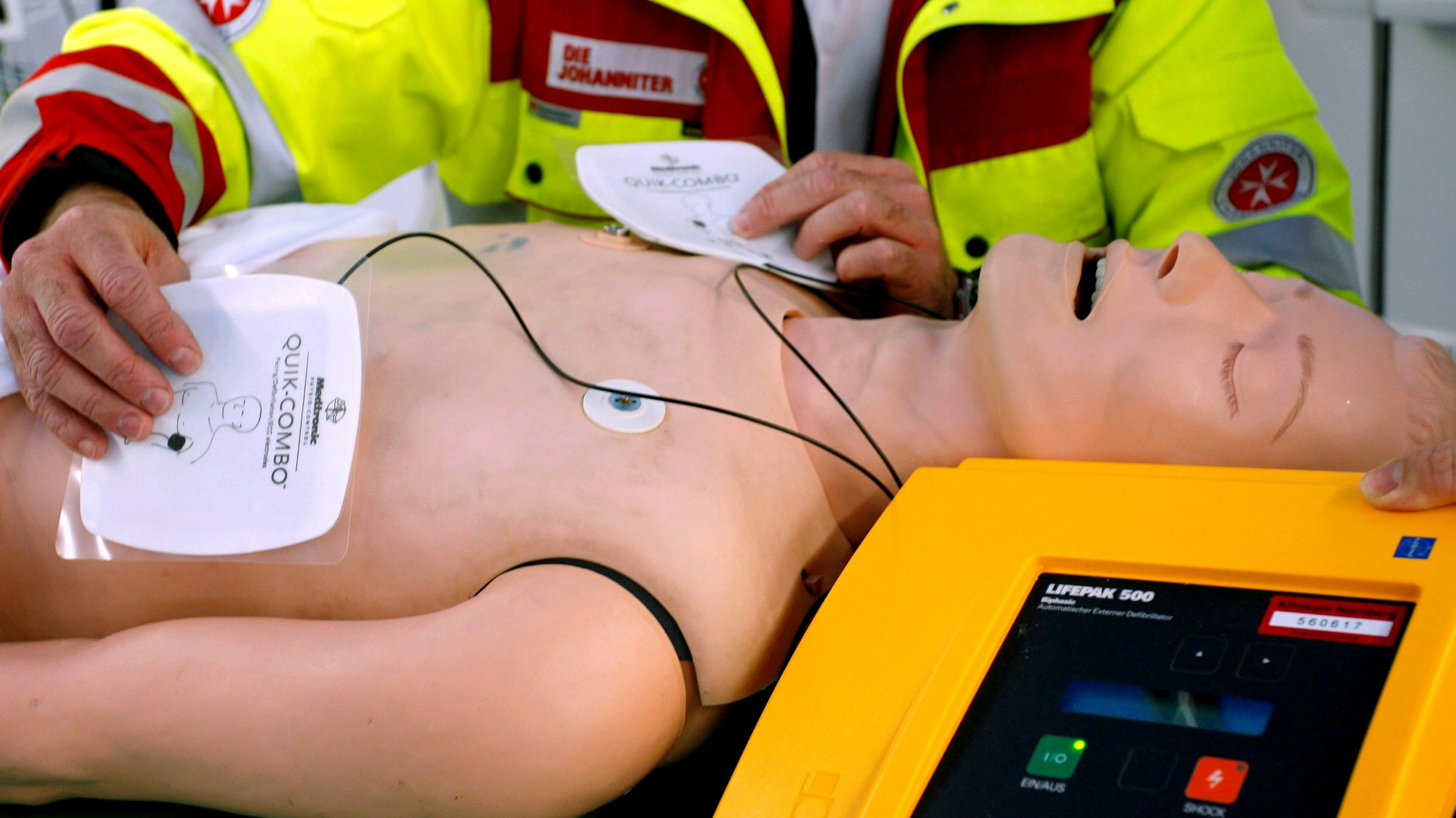 Ein Rettungssanitäter präsentiert einen Defibrillator an einer Puppe, die Elektroden-Pads liegen auf dem Torso.