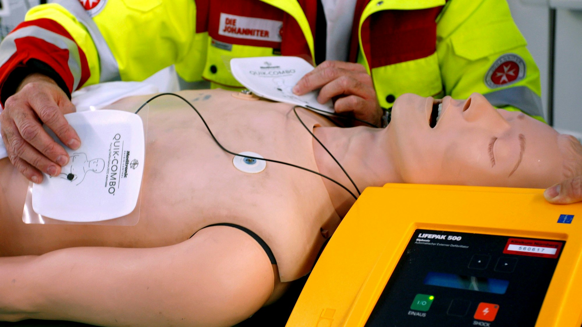 Ein Rettungssanitäter präsentiert einen Defibrillator an einer Puppe, die Elektroden-Pads liegen auf dem Torso.