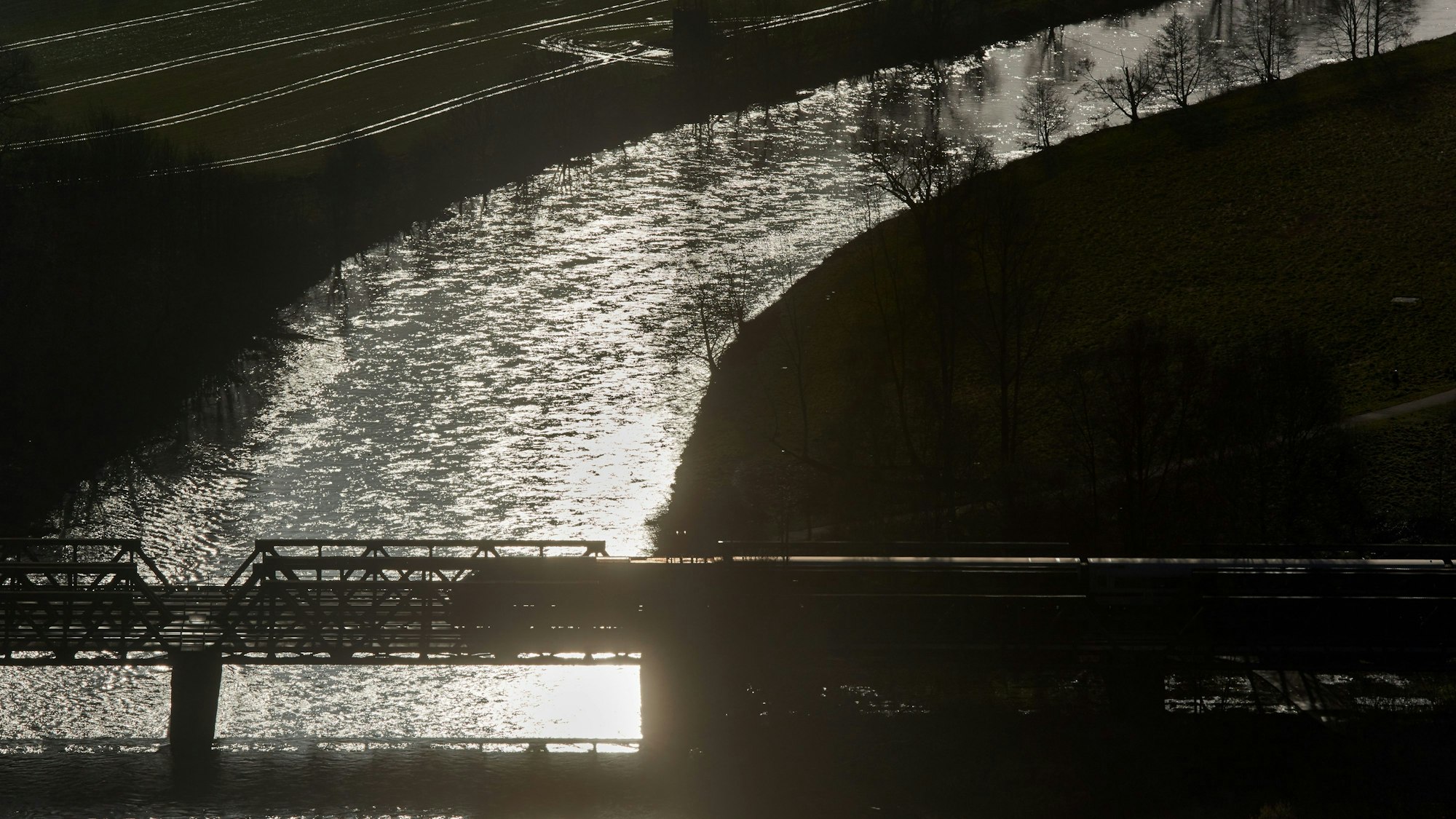 Ein Zug fährt im Gegenlicht über eine Brücke über das Flüsschen Lenne, in dessen Wellen sich die Morgensonne spiegelt.