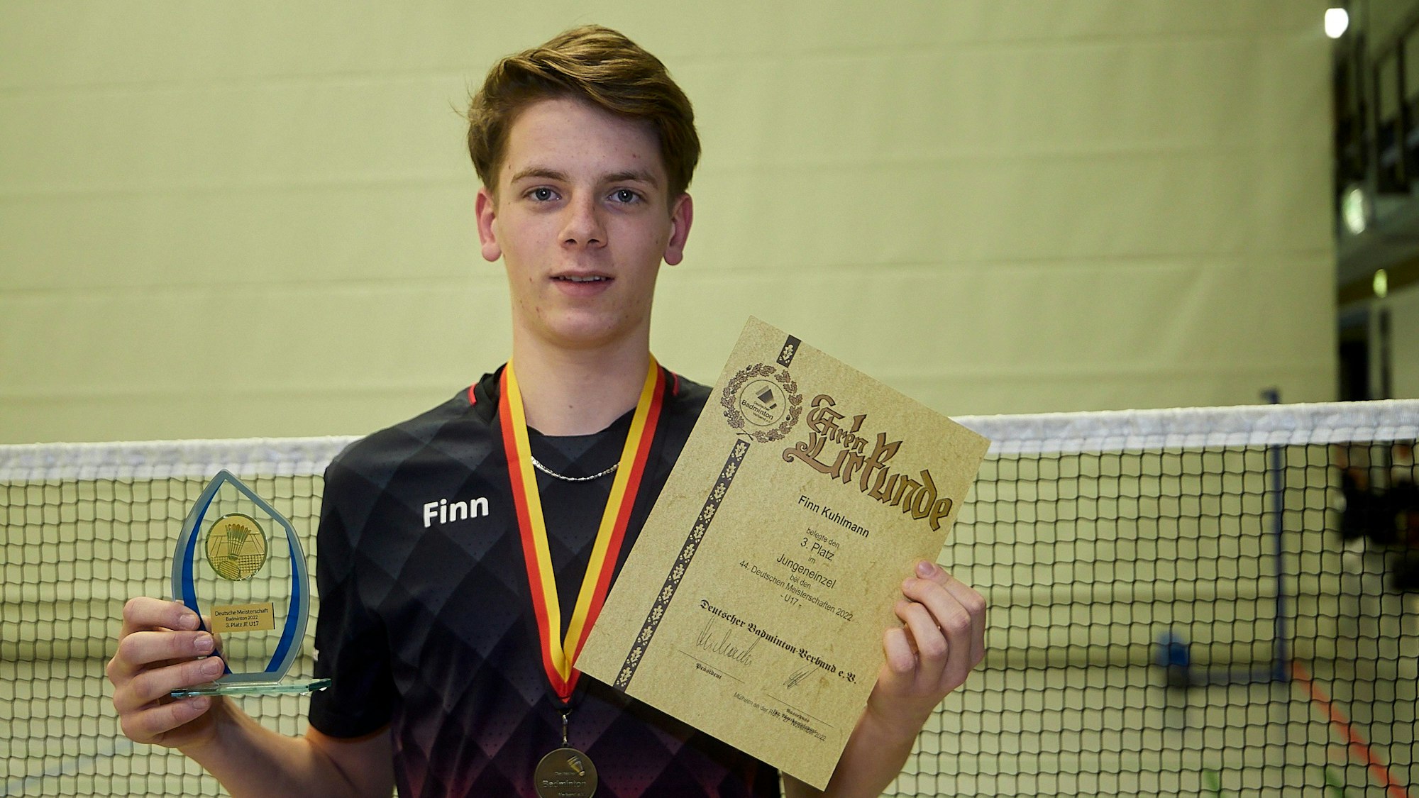 Badmintonspieler Finn Kuhlmann zeigt eine Urkunde und einen gläsernen Pokal.