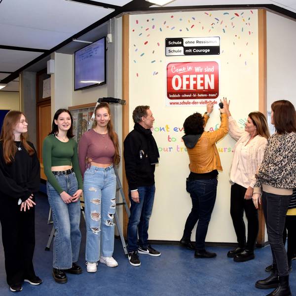 Schülerinnen und Lehrkräfte stehen vor einem Schild in einer Schule.