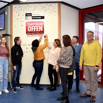 Schülerinnen und Lehrkräfte stehen vor einem Schild in einer Schule.
